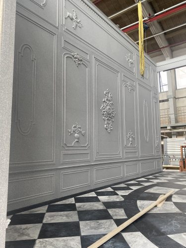 Eine reich verzierte graue Wand mit barocken Ornamenten steht auf einem Schachbrettboden in einer Werkhalle.