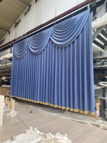 Blauer Bühnenvorhang mit goldener Borte hängt in einer Werkstatt mit verschiedenen Materialien im Hintergrund.