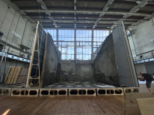 Bühnenbild in einer Halle mit schrägen, verwitterten Wänden und schwarz-weiß-kariertem Boden, eine Person steht rechts.