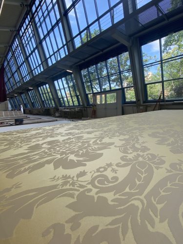 Große Halle mit hohen Fenstern, Tageslicht fällt auf eine große helle Fläche mit floralem Muster im Vordergrund.
