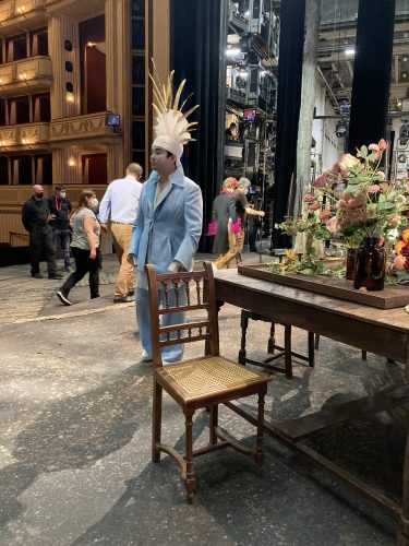 Person in hellblauem Anzug und auffälligem Federkopfschmuck steht neben einem Tisch mit Blumenarrangement auf einer Theaterbühne.