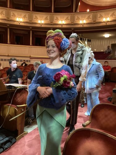 Eine Frau mit buntem Kopfschmuck und Blumenstrauß geht lächelnd durch einen Theatersaal, gefolgt von weiteren kostümierten Personen.