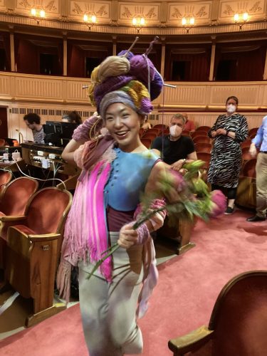 Eine Frau in farbenfrohem Kostüm mit auffälligem Kopfschmuck und Blumenstrauß steht lächelnd im Zuschauerraum eines Theaters.