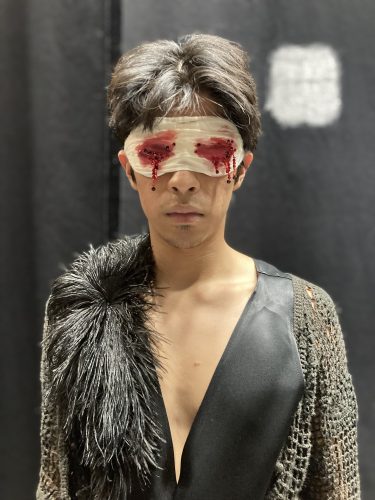 Eine Person mit verbundenen Augen, auf deren Binde rote, blutähnliche Flecken zu sehen sind, trägt ein auffälliges, schwarz-graues Outfit.