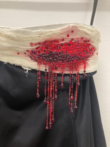 Weißer Stoff mit roten Perlen und Pailletten, die wie Blut aussehen und an einem schwarzen Rock herunterhängen.