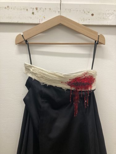 Schwarzes Kleid auf einem Holzbügel mit weißem, ausgefranstem Stoff am Ausschnitt und roten Perlen, die wie Bluttropfen wirken.