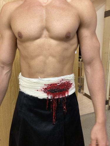 Ein muskulöser Oberkörper mit einem weißen Verband um die Taille, der blutbefleckt ist und Blut heruntertropft.