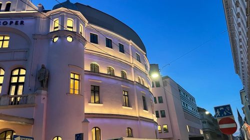 Historisches Gebäude mit abgerundeter Fassade und beleuchteten Fenstern bei Abenddämmerung in einer Stadtstraße.