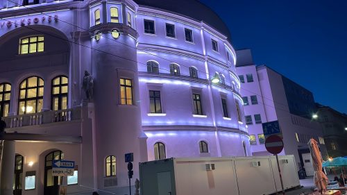 Ein historisches Gebäude mit lila Beleuchtung am Abend, davor Baustellencontainer und Baustellenschilder auf der Straße.