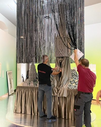 Zwei Männer dekorieren eine Installation mit langen schwarzen Fransen und silbernen Stoffen, darunter steht eine Statue mit Schlossmotiven.