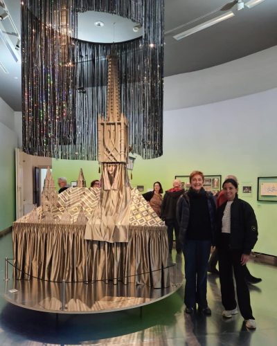 Zwei lächelnde Personen stehen vor einem silberfarbenen Architekturmodell, umgeben von weiteren Besuchern in einem Ausstellungsraum.