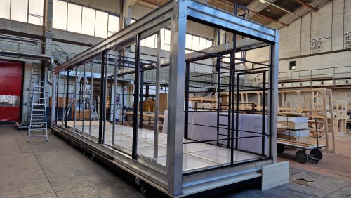 Metallrahmen eines großen rechteckigen Containers in einer Werkshalle, mit Leitern und Baumaterialien im Hintergrund.