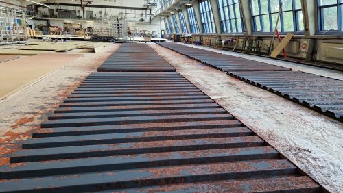 Mehrere lange, schwarz gestrichene Metallteile liegen nebeneinander in einer großen Industriehalle mit hohen Fenstern.