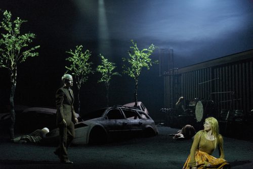 Mehrere Menschen auf einer düsteren Theaterbühne mit Schrottautos und jungen Bäumen; im Vordergrund sitzt eine Frau.