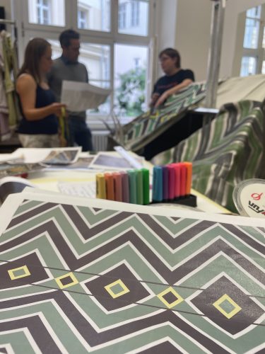 Nahaufnahme von bunten Stoffmustern mit geometrischen Mustern auf einem Tisch, im Hintergrund drei Personen in Diskussion.