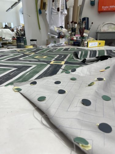 Stoffe mit geometrischen Mustern liegen auf einem Arbeitstisch in einem Atelier, umgeben von Pinseln und anderen Arbeitsmaterialien.