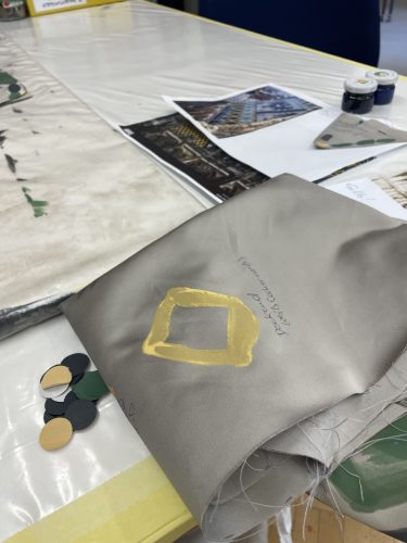 Grauer Stoff mit gelbem Quadrat, daneben Papierschnipsel, Fotos und Malutensilien auf einem Tisch.