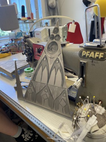 Ein dreieckiges, gezeichnetes Design mit skizzierten Mustern steht auf einem Tisch neben einer Nähmaschine und Nähzubehör.