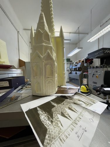 Ein Modell eines gotischen Turms steht auf einem Schreibtisch, umgeben von Skizzen und Fotos des Originalgebäudes in einem Arbeitsraum.