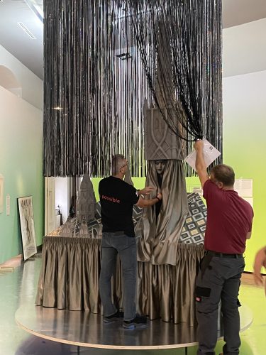 Zwei Männer arrangieren eine Installation mit schwarzen Lamellen und Stoff, während sie an einem Ausstellungsobjekt in einem Museum arbeiten.