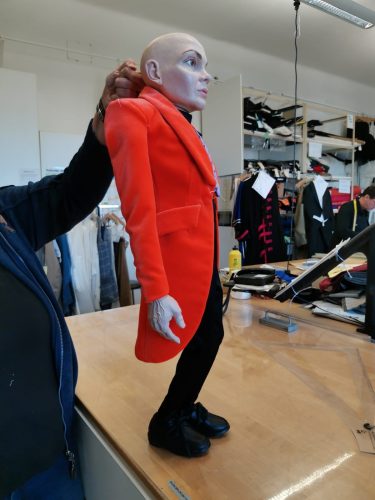 Eine Person hält eine Puppe mit Glatze und orangenem Mantel auf einem Holztisch in einem Raum voller Kleidung und Arbeitsmaterialien.