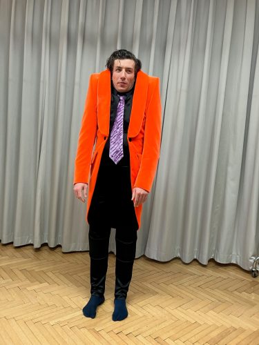 Ein Mann mit hochgezogener Schulterpartie trägt ein leuchtend oranges Sakko und eine gemusterte Krawatte, steht barfuß vor Vorhängen.
