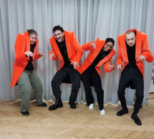 Vier Personen tragen leuchtend orange Jacken mit gepolsterten Schultern und stehen in lustigen, gebückten Posen vor einem grauen Vorhang.