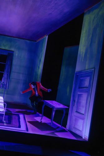 Eine Person in rotem Sakko lehnt in einem surreal beleuchteten, schrägen Zimmer mit violettem und grünem Licht.