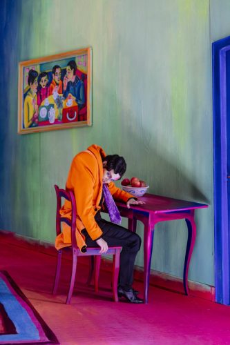 Ein Mann im orangefarbenen Mantel sitzt erschöpft an einem Tisch mit Obstschale in einem farbenfrohen, surrealen Raum.