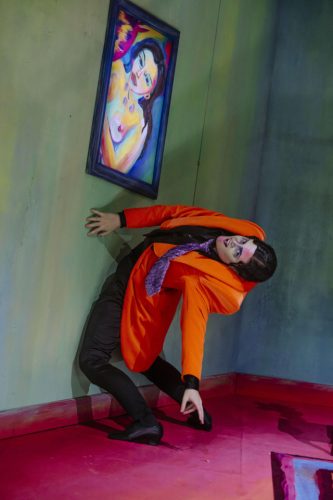 Eine Person im orangefarbenen Anzug lehnt sich ungewöhnlich gegen eine Wand mit einem farbenfrohen Gemälde einer Frau.