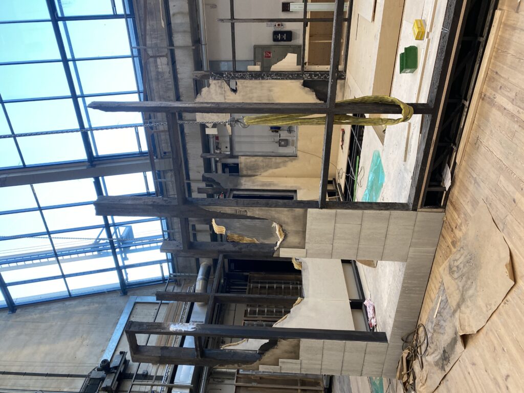 Teilweise zerstörte Theaterkulisse mit eingerissenen Wänden und freiliegenden Metallstreben in einer großen Halle.