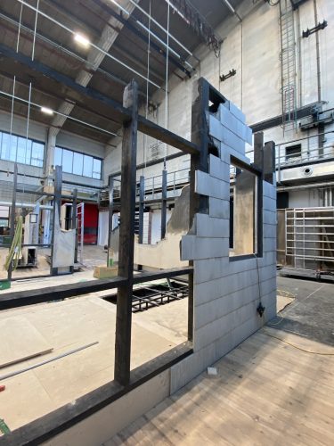 Teilweise errichtete Wand aus grauen Blöcken und dunklem Rahmen in einer großen Industriehalle.