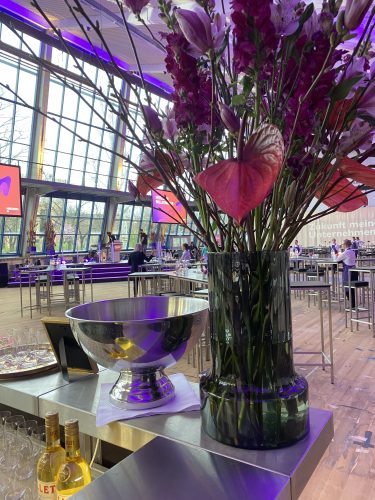 Großer Strauß aus rosa und lila Blumen in einer Vase auf einer Bar, daneben eine große Metallschale und einige Weinflaschen, im Hintergrund ein moderner Veranstaltungssaal.