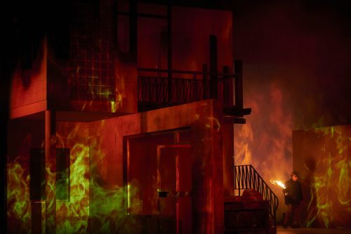 Eine Bühnenkulisse eines brennenden Hauses mit dramatischer Feuerprojektion und einer Person, die eine offene Flamme hält.