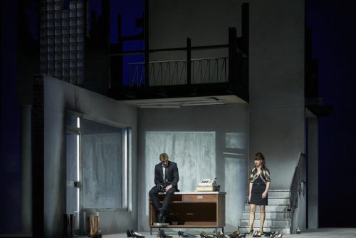 Zwei Personen in formeller Kleidung stehen in einer dramatisch beleuchteten, modernen Bühnenkulisse mit Schreibtisch, Treppe und verstreuten Schuhen.