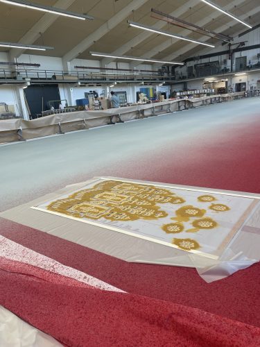 Ein großes Halleninnere mit rotem und grauem Boden, im Vordergrund eine Schablone mit goldener Farbe und Schneeflockenmotiven.