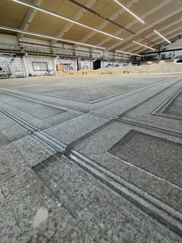 Große Halle mit einem weitläufigen, gemusterten Boden und hohen Deckenlampen, im Hintergrund sind Netze und Arbeitsbereiche zu sehen.