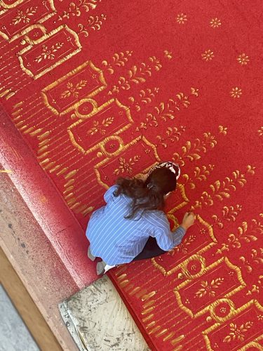 Ein Kind sitzt auf einem roten Teppich mit goldenen Mustern und schaut nach unten.
