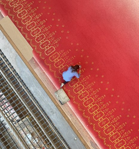 Eine Person mit langen Haaren kniet auf einem roten Teppich mit goldenen Mustern neben einem Metallgeländer.