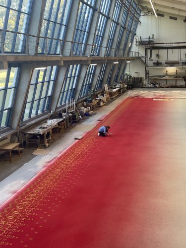 Eine Person kniet auf einem großen, rot gemusterten Teppich in einer hellen Werkhalle mit hohen Fenstern.