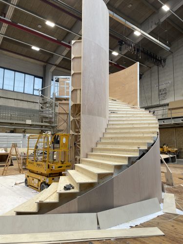 Große, unfertige Wendeltreppe aus Holz in einer Werkhalle mit Baugeräten und Leitern im Hintergrund.
