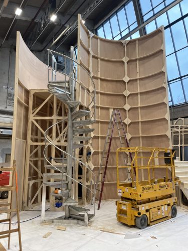 Eine große, halbkreisförmige Holzkonstruktion mit einer Wendeltreppe und einer gelben Hebebühne in einer Werkhalle.