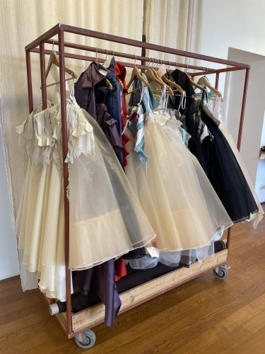 Kleiderständer mit mehreren eleganten Kleidern in verschiedenen Farben, hauptsächlich weiße, schwarze und lilafarbene Tüllkleider auf Holzkleiderbügeln.
