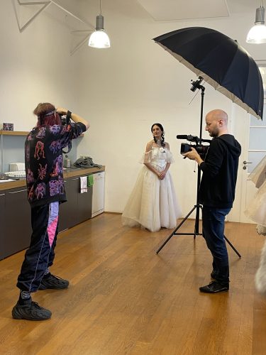 Eine Frau in einem weißen Kleid posiert für ein Fotoshooting, während zwei Personen sie fotografieren und filmen.