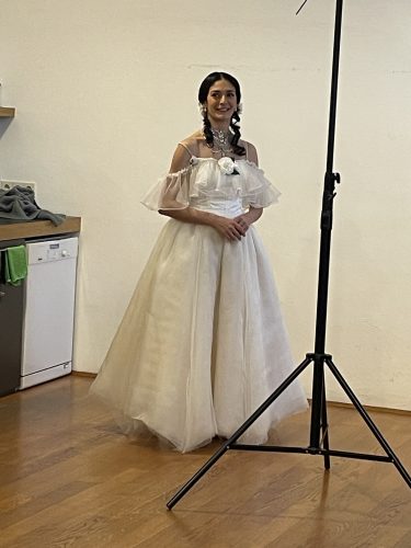 Frau in einem weißen, schulterfreien Ballkleid steht lächelnd in einem Raum mit hölzernem Boden neben einem Fotostativ.