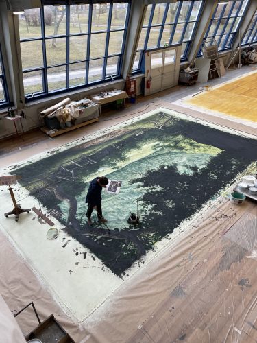 Eine Person malt ein großes Landschaftsgemälde auf einer riesigen Leinwand in einem lichtdurchfluteten Atelier.