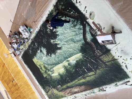 Eine Person malt ein großes Wandbild auf dem Boden, das eine idyllische Wald- und Wiesenlandschaft mit Zaun und Bäumen zeigt.