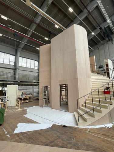 Holzkonstruktion eines zweistöckigen Gebäudes mit Treppe in einer Werkhalle, umgeben von Bauwerkzeugen und Materialien.