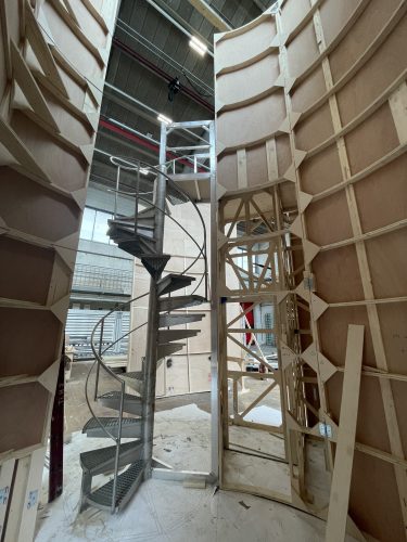 Wendeltreppe aus Metall inmitten eines unfertigen Holzbaus mit sichtbaren Streben und Baustellenatmosphäre.