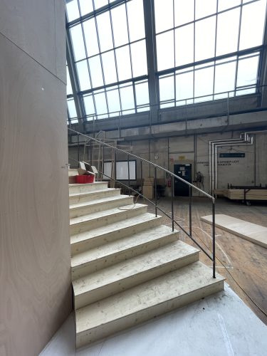 Eine helle Werkhalle mit großen Fenstern, einer geländerlosen Holztreppe und einem roten Behälter auf der Treppe.
